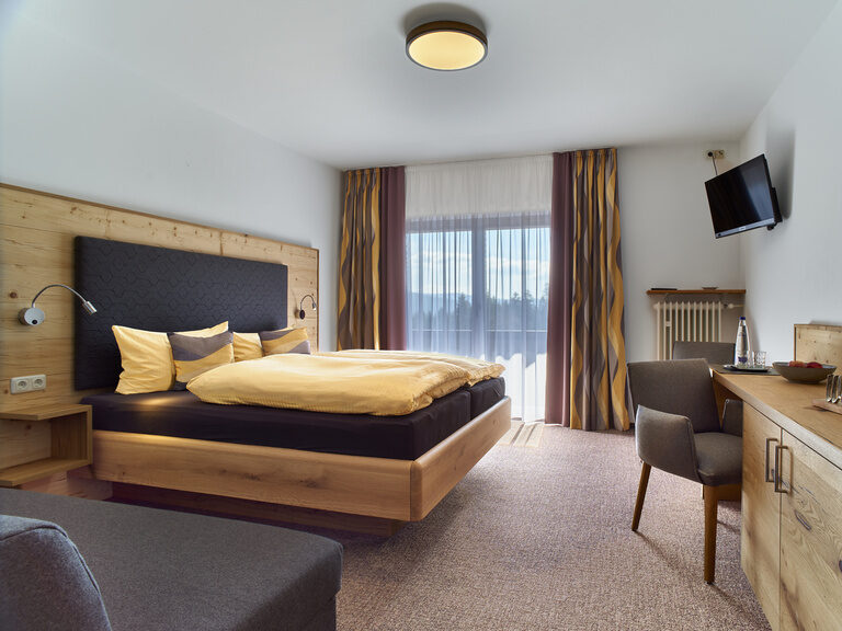 Ein komfortables Schlafzimmer mit weitem Balkonfenster und wohnlicher Holzeinrichtung in der Pension Kilger.